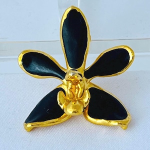 May include: A gold-toned metal flower brooch with black enamel petals. The brooch has a detailed, three-dimensional design, with a gold center and edges. The flower is a stylized orchid, perfect for adding a touch of elegance to any outfit.