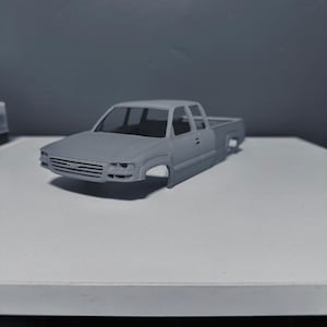 May include: A gray model pickup truck with a crew cab design. The truck is missing its wheels and sits on a white surface. The front grille and headlights are visible, and the truck bed is empty.