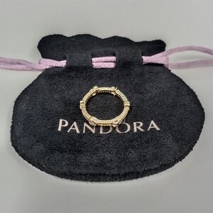 May include: A gold-colored ring with small, clear stones is displayed on a black pouch with the word "PANDORA" printed in white. The ring has a repeating design and is centered on the pouch.