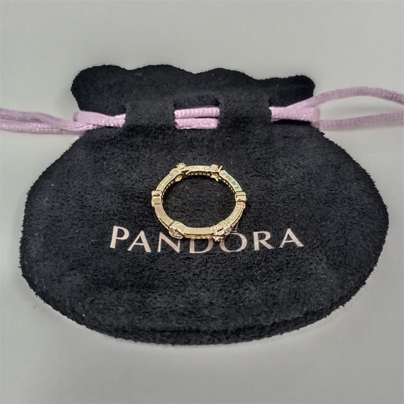 May include: A gold-colored ring with small, clear stones is displayed on a black pouch with the word "PANDORA" printed in white. The ring has a repeating design and is centered on the pouch.