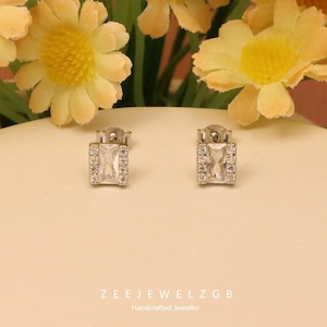 May include: Two silver stud earrings, each featuring a rectangular, clear central stone, framed by smaller, clear stones. The earrings are set against a cream background, with yellow flowers above. The text "ZEEJEWELZGB Handcrafted Jeweller" is visible at the bottom.