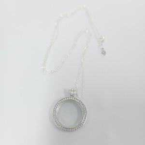 Puede incluir: Un collar con colgante de medallón plateado y cadena fina. El medallón redondo está adornado con pequeñas piedras transparentes en el borde. El collar se presenta sobre un fondo blanco, resaltando el diseño y los detalles de la joya.