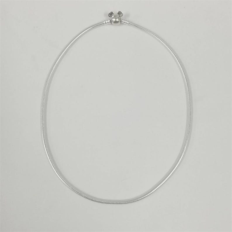 May include: A silver-colored necklace with a round, snake-like chain. The clasp features a small, decorative design. The necklace is displayed against a plain white background, highlighting its simple elegance.