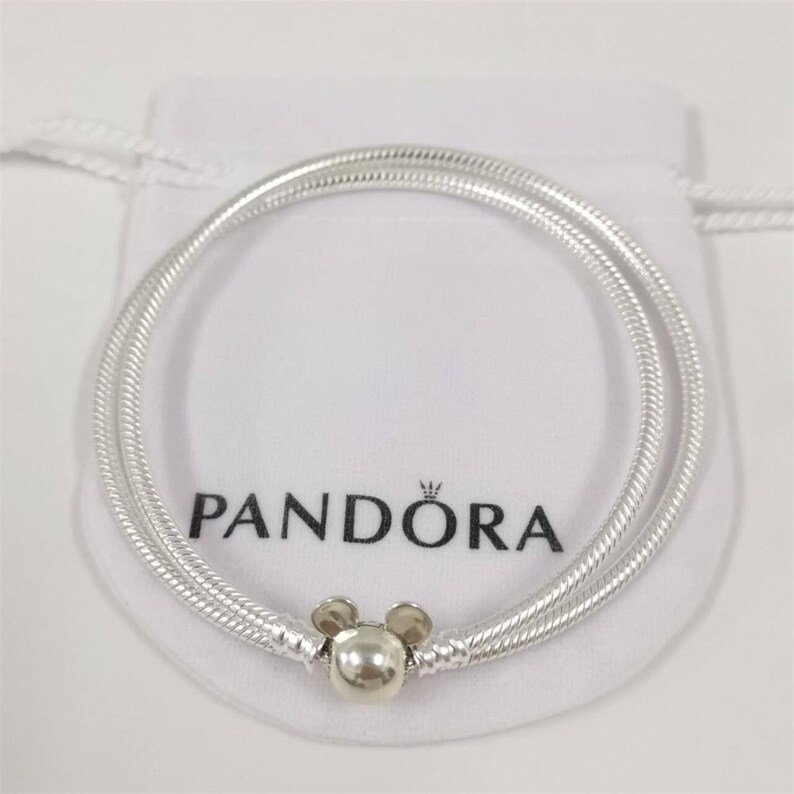 May include: A silver Pandora bracelet with a Mickey Mouse charm. The bracelet is coiled and rests on a white pouch with the Pandora logo printed in black. The charm is a silver sphere with two small silver circles on top.