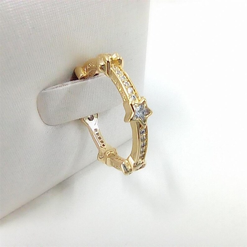 May include: A gold-colored ring with a star-shaped design and small, clear gemstones. The ring is set against a white background, showcasing its intricate details and craftsmanship. The ring is a piece of jewelry.
