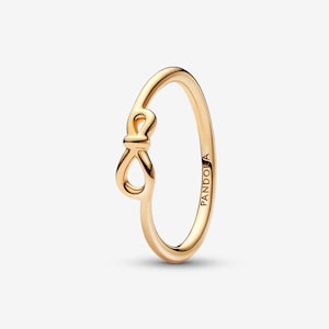 Puede incluir: Un anillo de tono oro rosa pulido que presenta un delicado diseño de nudo inspirado en el infinito en la banda. La superficie metálica es lisa y reflectante, con el nombre de la marca PANDORA grabado en letras mayúsculas en el interior.