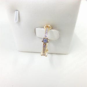 May include: A gold-colored ring with a star-shaped blue gemstone and clear, small gemstones. The ring is displayed on a white surface. The ring's design features a star in the center and smaller stones along the band.