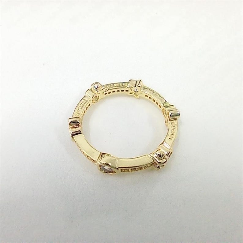 May include: A gold-colored ring with a repeating geometric design. The ring features small, clear gemstones and is set against a white background. The word "elegance" is visible on the ring.