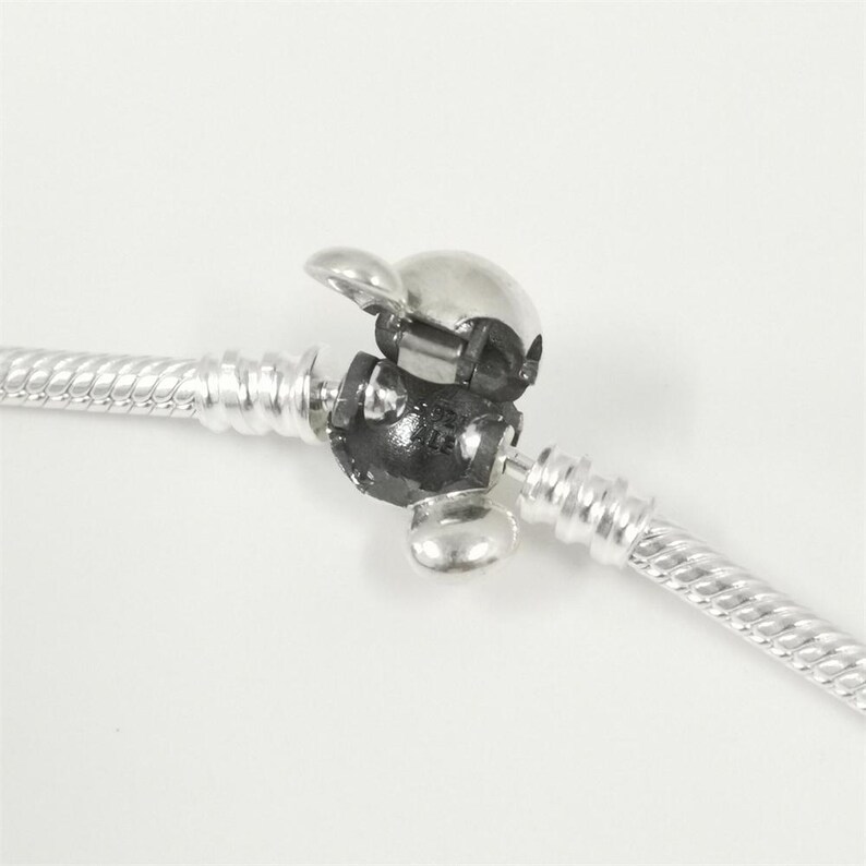 May include: A silver-colored bracelet clasp with a hinged, rounded top and a black base. The clasp is attached to a silver, twisted rope-style bracelet. The background is plain white.