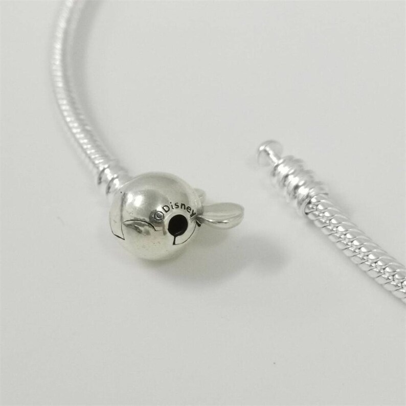 May include: A silver bracelet with a Mickey Mouse-shaped clasp. The clasp is detailed with the word "Disney" and a small black circle. The bracelet is unclasped, showing the two ends of the chain.