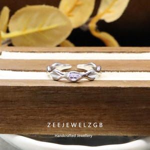 May include: A silver ring with an open design, featuring a central oval, light purple gemstone. The ring has a floral or leaf-like pattern on the band. The text "ZEEJEWELZGB" and "Handcrafted Jewellery" are visible. The ring is displayed on a wooden surface.