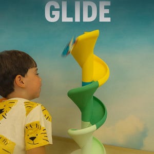Puede incluir: Un niño observa un juguete colorido con el texto "GRAVITY GLIDE" arriba. El juguete presenta un diseño de tobogán en espiral amarillo y verde. El niño lleva una camiseta blanca con un estampado de cara de tigre amarillo.