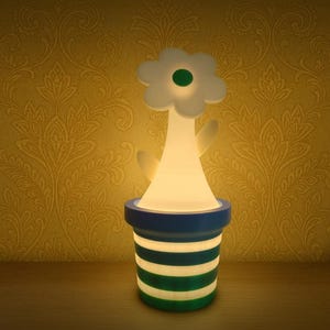 May include: A flower-shaped night light with a white flower head and a green center. The light sits in a blue and green striped pot. The lamp is illuminated, casting a warm glow.