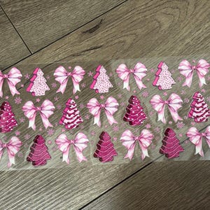 May include: A sheet of Christmas-themed stickers featuring pink bows and Christmas trees. The bows have white accents, and the trees are decorated with various patterns in shades of pink. The stickers are arranged in rows.