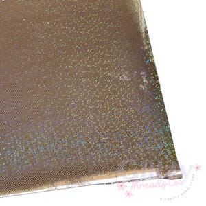 May include: Close-up of a sheet of shimmering, gold-colored fabric with a subtle, dotted pattern. The material has a reflective quality, catching light and displaying iridescent hues. The edge of the fabric is visible, suggesting it's a cut piece, possibly for crafting.