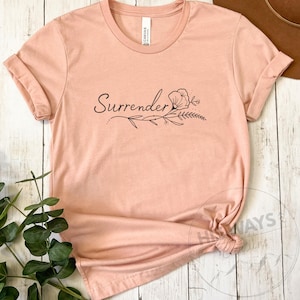May include: A light pink t-shirt with rolled-up sleeves. The word "Surrender" is printed in a cursive font, accompanied by a floral design. The shirt is knotted at the bottom.