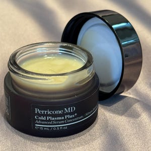 May include: An open jar of Perricone MD Cold Plasma Plus+ Advanced Serum Concentrate. The jar is dark brown with a clear glass top, revealing a yellow serum. The black lid is next to the jar. The text on the jar reads "15 mL / 0.5 fl oz."