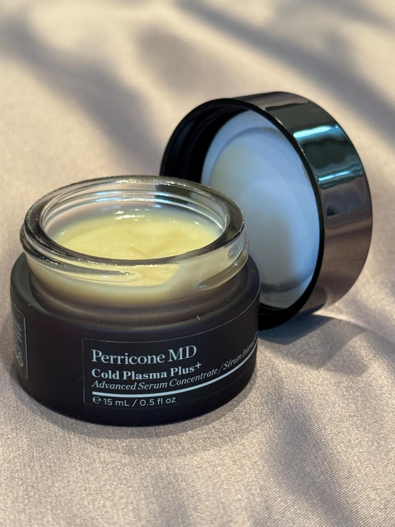 May include: An open jar of Perricone MD Cold Plasma Plus+ Advanced Serum Concentrate. The jar is dark brown with a clear glass top, revealing a yellow serum. The black lid is next to the jar. The text on the jar reads "15 mL / 0.5 fl oz."