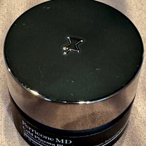May include: A black jar with a silver rim and a black lid. The lid has a silver star-shaped emblem. The jar contains Perricone MD Cold Plasma Plus+ Advanced Serum Concentrate. The jar is 0.5 fl oz.
