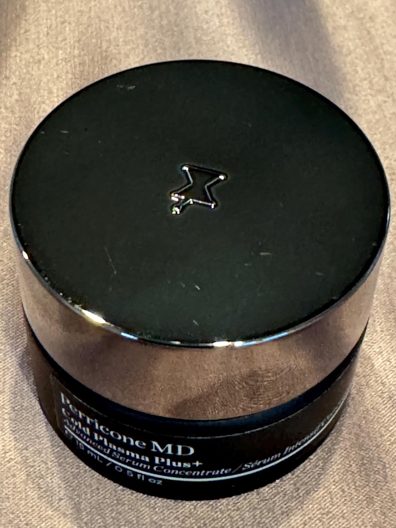 May include: A black jar with a silver rim and a black lid. The lid has a silver star-shaped emblem. The jar contains Perricone MD Cold Plasma Plus+ Advanced Serum Concentrate. The jar is 0.5 fl oz.