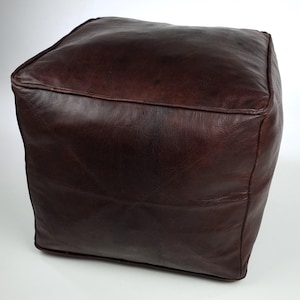 Puede incluir: Un puf cuadrado de cuero marrón oscuro. El puf tiene una superficie texturizada y está hecho de una sola pieza de cuero. Las costuras son visibles en la parte superior y los lados. El puf tiene forma de cubo.