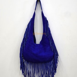 Könnte beinhalten: Eine leuchtend blaue Wildleder-Umhängetasche mit langen Fransen. Die Tasche hat eine abgerundete Form und lange Riemen und hängt an einer weißen Wand. Die Fransen hängen von der Unterseite und den Seiten der Tasche.