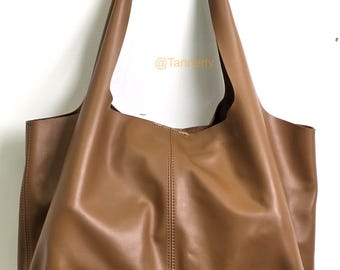 May include: A large, tan leather tote bag with a single handle and long shoulder straps. The bag has a simple, minimalist design with a smooth texture and a spacious interior. The bag is shown against a white background.