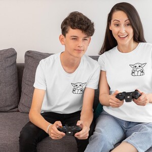May include: Two people wearing white t-shirts with a black and white graphic of a character. They are sitting on a gray couch and holding black game controllers. The t-shirts are a crew neck style.