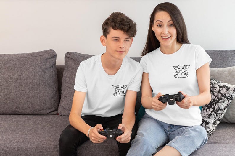 May include: Two people wearing white t-shirts with a black and white graphic of a character. They are sitting on a gray couch and holding black game controllers. The t-shirts are a crew neck style.