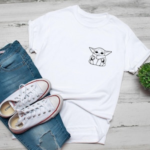 May include: White t-shirt with a black and white graphic of a cartoon character. The shirt is paired with blue jeans and white sneakers with red accents. A small green plant is also in the image.
