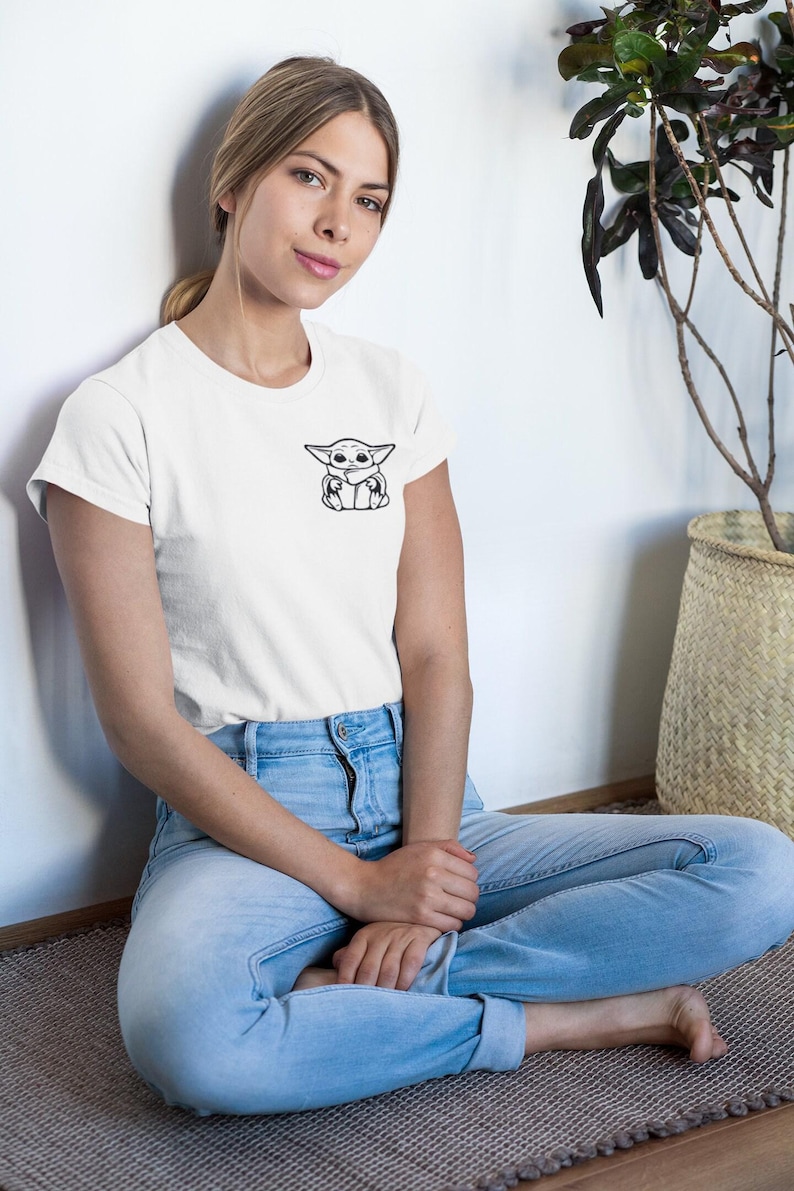 May include: White t-shirt featuring a black and white graphic of a character. The t-shirt is made of a soft material and has short sleeves. The person is wearing blue jeans.
