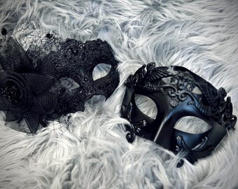 Puede incluir: Dos máscaras negras de mascarada. Una máscara presenta un encaje intrincado y un acento floral con un velo. La otra máscara tiene un diseño decorativo con un águila y volutas. Ambas máscaras están sobre un fondo gris y esponjoso.