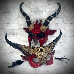 Può includere: Due maschere a tema diavolo. La maschera superiore è rossa con corna nere avvolte in spago. La maschera inferiore è rossa con filigrana dorata, corna nere e piume nere. Entrambe le maschere hanno un aspetto strutturato e sono per costumi.