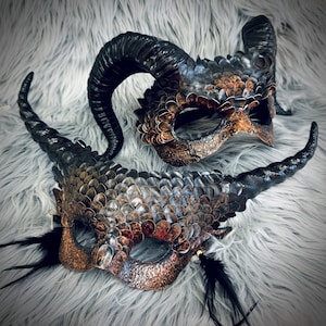 May include: Two ornate, handcrafted masks with a fantasy theme. The masks feature textured, scale-like patterns in shades of bronze, copper, and black. Each mask has large, curved horns and is embellished with black feathers.