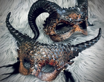 Könnte beinhalten: Zwei kunstvoll gefertigte Masken mit Fantasy-Thema. Die Masken weisen strukturierte, schuppenartige Muster in Bronze-, Kupfer- und Schwarztönen auf. Jede Maske hat große, gebogene Hörner und ist mit schwarzen Federn verziert.