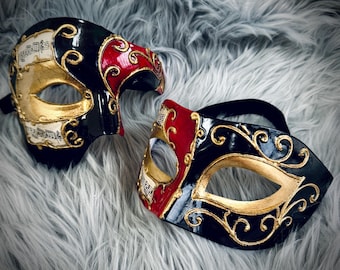 May include: Two Venetian masks. One mask is black with gold accents and musical notes, and the other is red and black with gold accents. Both masks have a gold eye opening and are set against a gray, furry background.