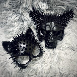 Puede incluir: Dos máscaras negras con detalles de púas. Una máscara tiene forma de gato, la otra es una cara de lobo o oso. Ambas máscaras tienen un acabado brillante y están sobre un fondo gris y esponjoso.