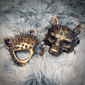 May include: Two ornate, gold-colored masks. One is cat-shaped with metal spikes, the other is a wolf or fox with detailed fur texture. Both masks are on a fluffy, light gray surface.