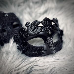 May include: A black masquerade mask with intricate details, including a winged creature and floral accents. The mask is set against a soft, textured background, creating a dramatic effect. The mask is designed for a formal event.