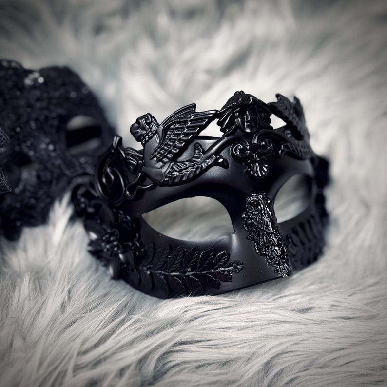 May include: A black masquerade mask with intricate details, including a winged creature and floral accents. The mask is set against a soft, textured background, creating a dramatic effect. The mask is designed for a formal event.