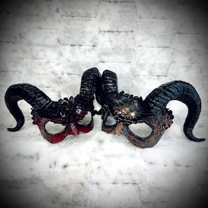 May include: Two horned masks with intricate details. One mask is primarily red and black, while the other is bronze and black. Both masks feature large, curved horns and decorative elements around the eye openings, creating a dramatic and fantasy-inspired aesthetic.