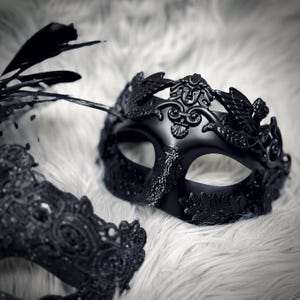 May include: Black masquerade mask with a decorative design. The mask features a detailed face with a crown of leaves and feathers. The mask is set against a white, fluffy background.