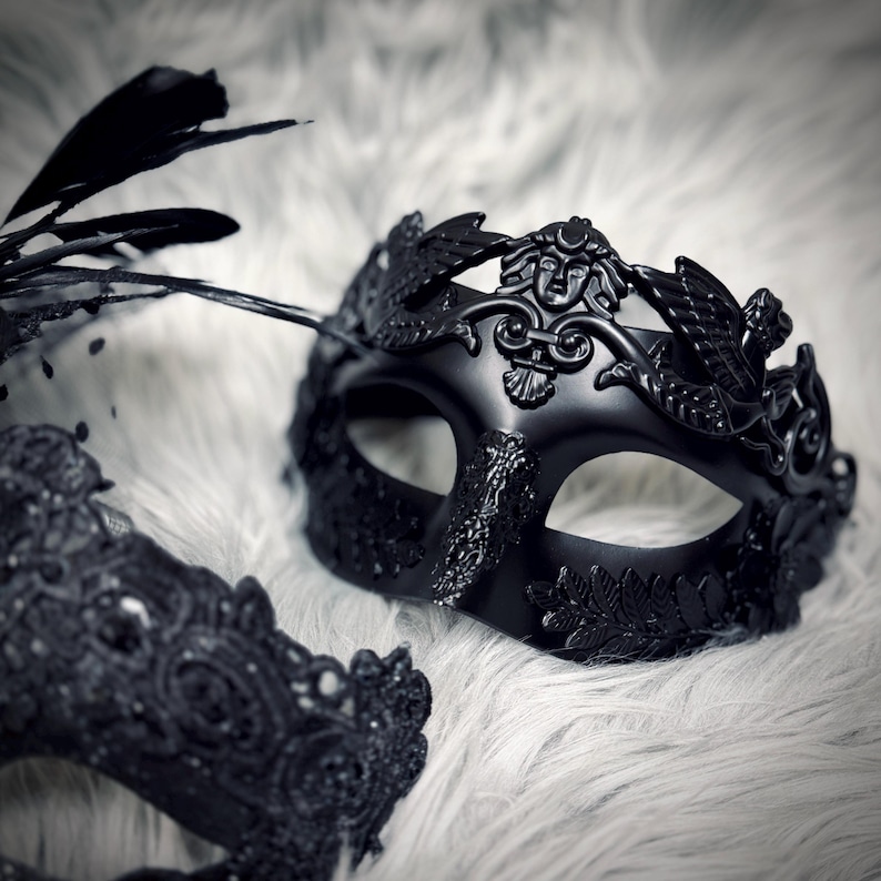 May include: Black masquerade mask with a decorative design. The mask features a detailed face with a crown of leaves and feathers. The mask is set against a white, fluffy background.