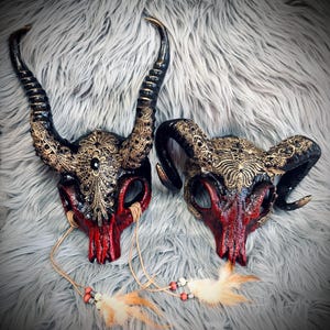 Può includere: Due maschere decorative a forma di teschio animale con lunghe corna nere ricurve e dettagli in filigrana dorata. Le superfici dei teschi presentano una sfumatura rossa e nera, arricchite da gemme nere, cordini in pelle e piume.