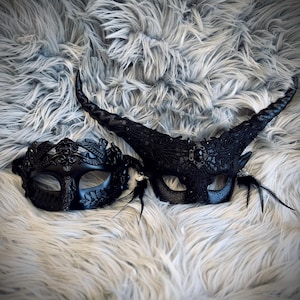 May include: Two black masquerade masks with intricate designs. One mask features a crown design, while the other has large horns and feather accents. Both masks are set against a fluffy, gray background.