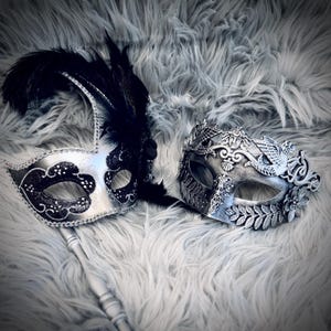 Pode incluir: Duas máscaras decorativas de carnaval. Uma é prateada com detalhes pretos e penas. A outra é prateada com desenhos florais e de folhas. Ambas as máscaras estão sobre uma superfície cinzenta e fofa.