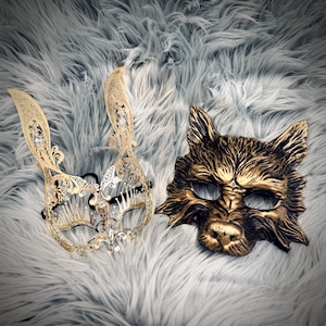 May include: Two decorative masks on a fluffy, gray surface. One is a gold-colored, filigree rabbit mask with long ears. The other is a gold-colored wolf mask with detailed fur texture. Both masks are designed for a masquerade or costume event.