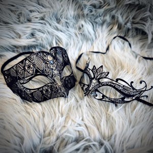 May include: Two black metal masquerade masks with intricate filigree designs. One mask features a geometric pattern, while the other has a floral motif. Both masks have eye openings and attached black ribbons.
