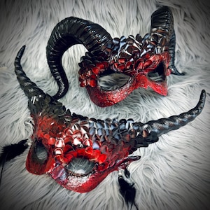 May include: Two ornate, handmade masks with horns. The masks are primarily red and black, with a scale-like texture. The horns are black and curved. Feathers are attached to the bottom mask. The masks are likely for a costume or masquerade.