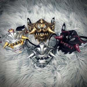 May include: Four detailed Oni masks in silver, gold, and black with red accents. Each mask features sharp teeth and horns, with black straps for wearing. The masks are displayed on a fluffy, white surface.